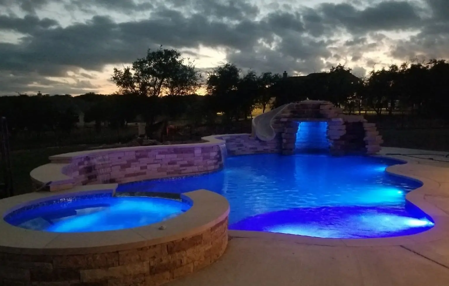 Pool and raised stone spa lit blue at dusk, with a rock grotto and slide