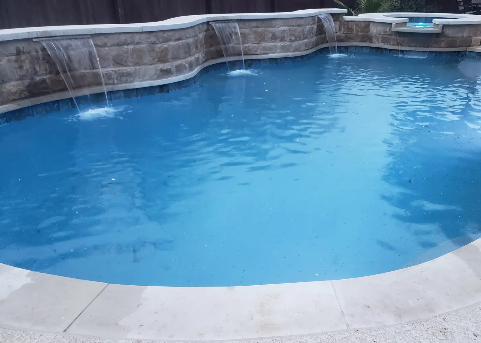 Freeform pool with three sheer waterfalls from a curved stone wall and a raised spa