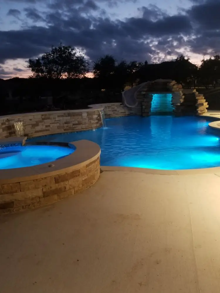 Pool with grotto waterfall and round spa lit blue at dusk